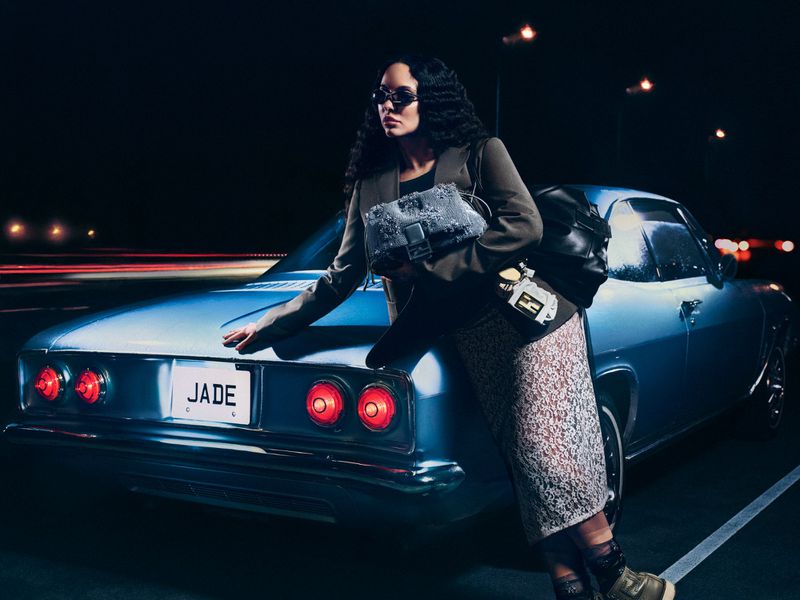 A woman in sunglasses and a jacket poses against a blue vintage car at night with motion blurred lights in the background.