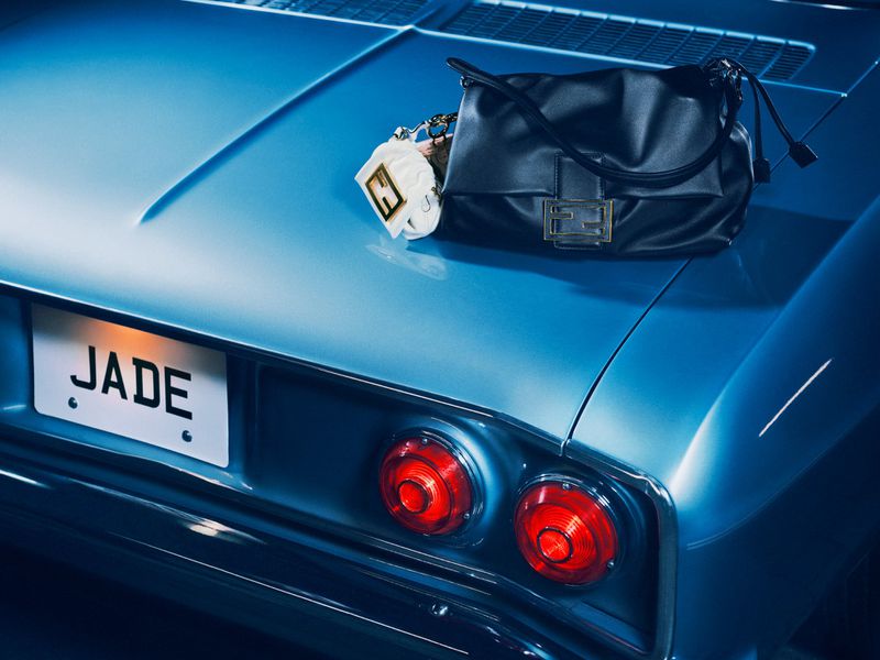 Blue vintage car with a black purse and keychain accessory on the trunk. The license plate reads 'JADE', and tail lights glow.