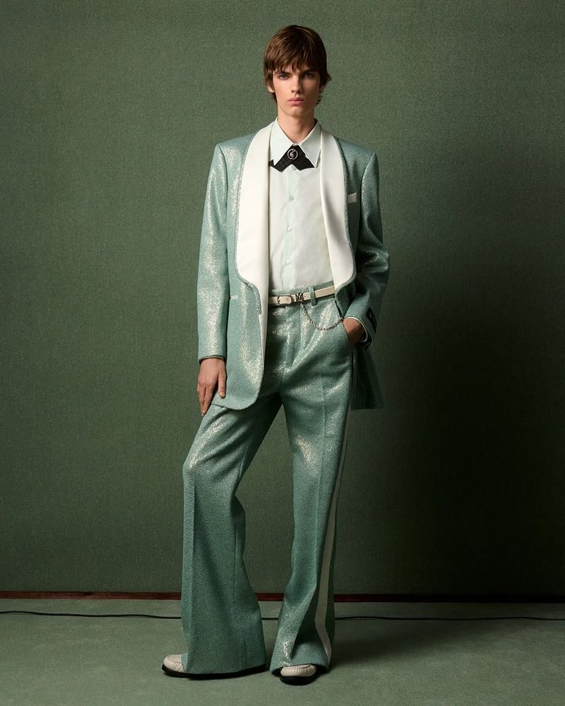 Fashionable man in a light blue suit with white accents posing against a green background, showcasing a retro-inspired style.