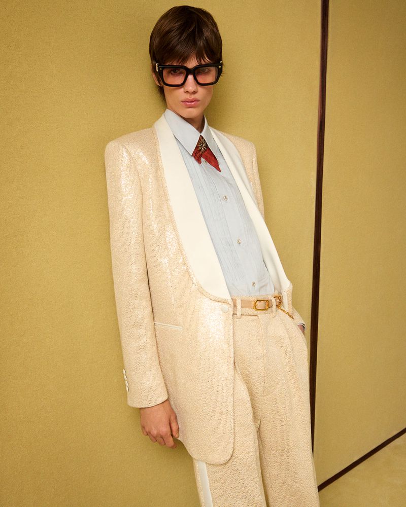 Adult model in a cream-colored sequin suit with white lapels, a button-down shirt, and dark glasses, posing against a cream backdrop.