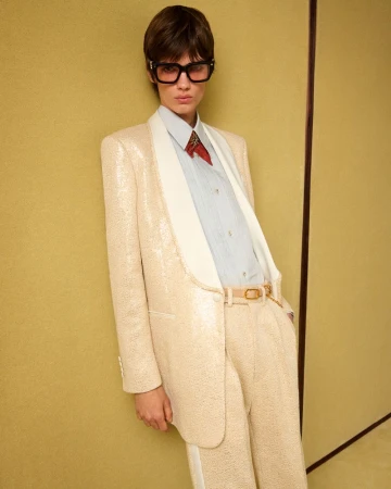 Adult model in a cream-colored sequin suit with white lapels, a button-down shirt, and dark glasses, posing against a cream backdrop.