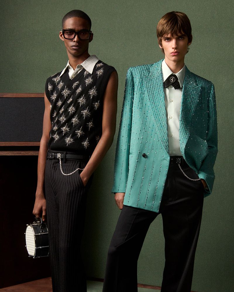 Two young male fashion models pose in coordinated suits against a green backdrop in a studio fashion editorial shot.