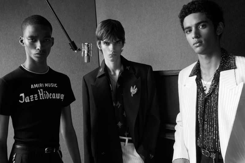 Three male models showcase Amiri Music fashion in a monochrome studio shot, featuring a vintage microphone and stylish clothing details.
