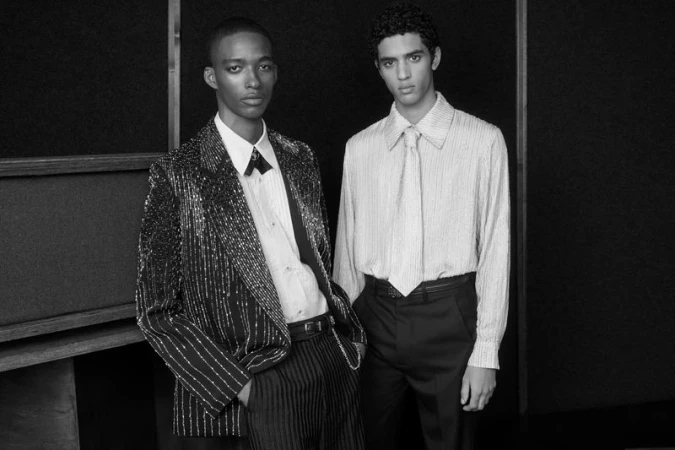 Two male models in tailored formal wear standing against a dark panelled wall for Amiri Spring/Summer 2025 campaign.