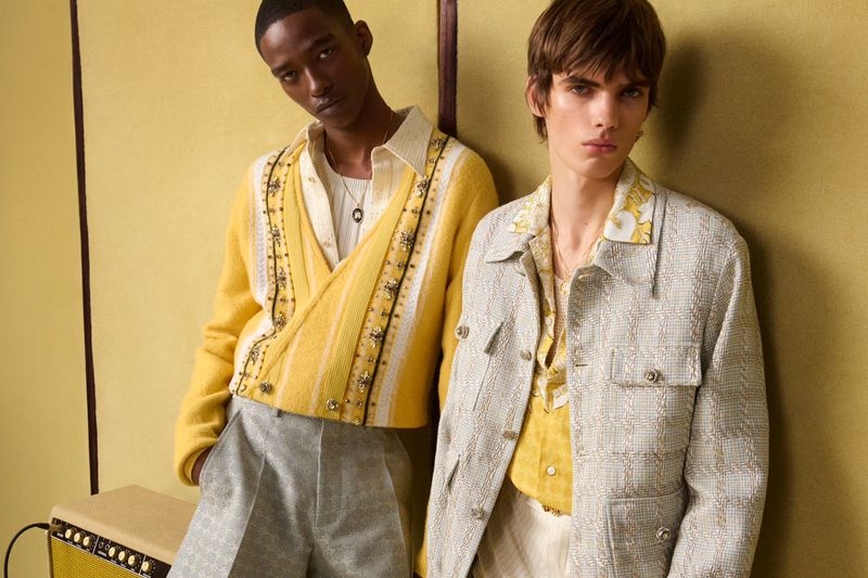 Two young adult models in patterned yellow cardigans and gray trousers pose against a yellow wall, showcasing stylish fashion.