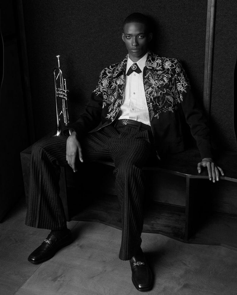 Black and white portrait of an adult male in an embroidered jacket sitting with a trumpet in a dimly lit indoor setting.