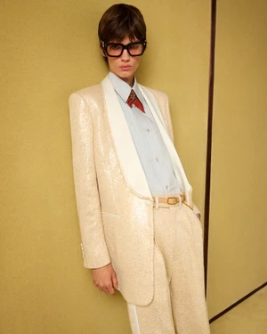 Model wearing a shimmering cream sequined suit and black sunglasses leaning against a textured yellow wall
