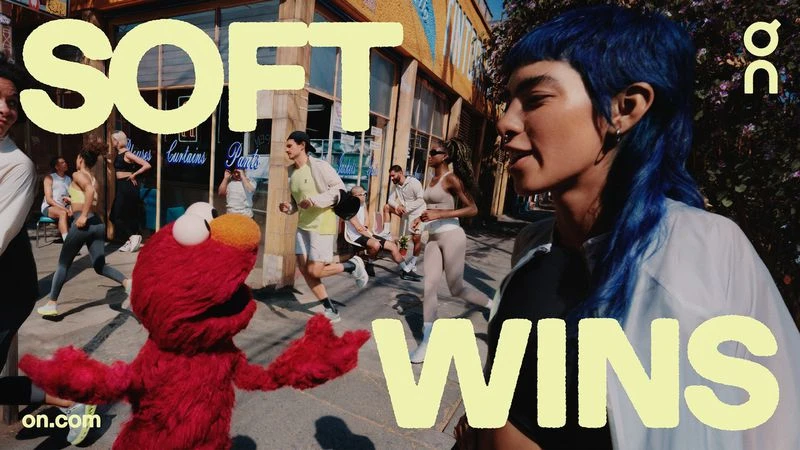 Outdoor shot of a diverse group of people running on a city street alongside a red Elmo character in front of storefronts.