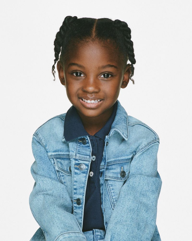 Portrait of a smiling young African-American girl with braided hair, wearing a light blue denim jacket and navy collared shirt, set against a white backdrop.