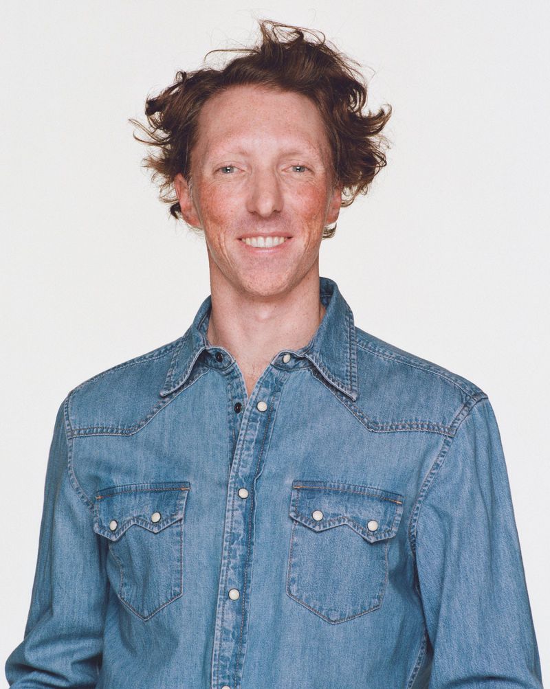 Portrait of a man with red hair and denim shirt against a plain white background, smiling and looking directly at the camera.