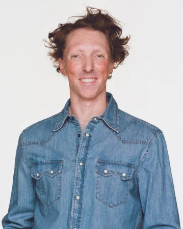 Portrait of a man with red hair and denim shirt against a plain white background, smiling and looking directly at the camera.