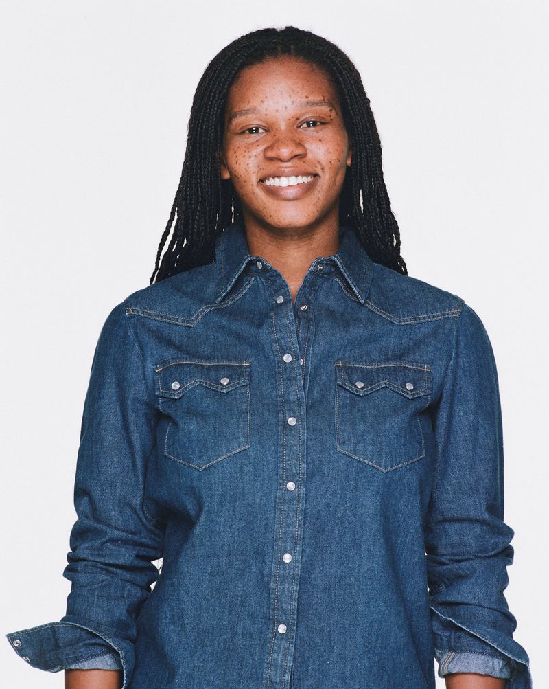 Portrait of a smiling Black woman with braided hair wearing a denim shirt against a white background in a studio setting.  