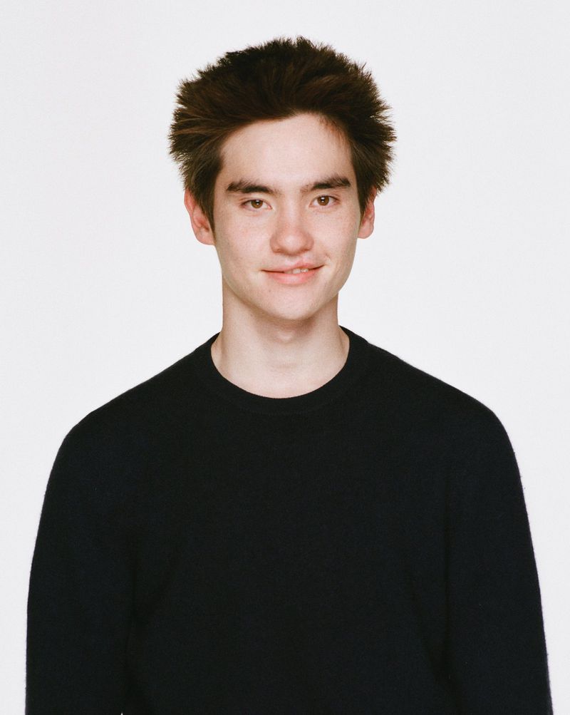 Close-up portrait of a young adult male with dark hair and a black sweater against a bright white backdrop in a studio setting.