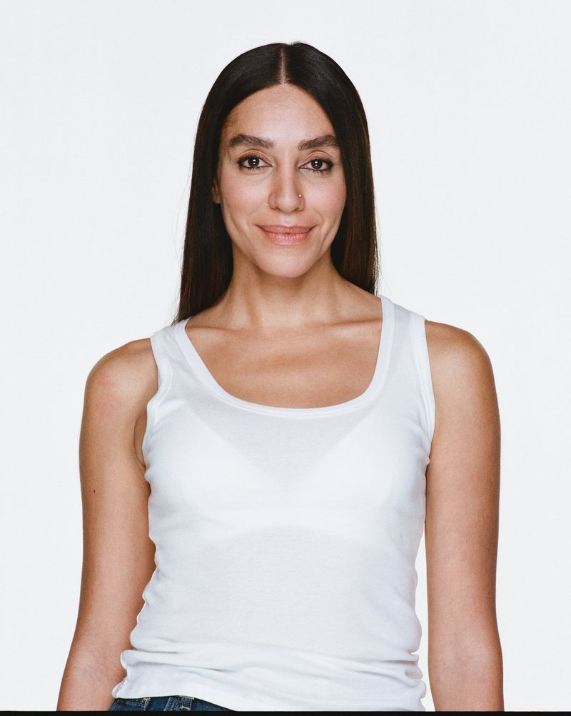 Portrait of adult woman with long brown hair smiling in white tank top against a white backdrop in a studio setting with eye-level angle.