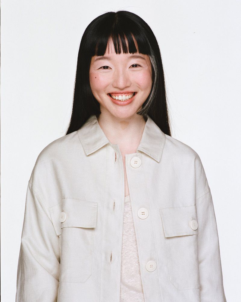 Smiling Asian woman in a buttoned shirt poses against a white backdrop, her black hair framing her face in a portrait-style photograph.