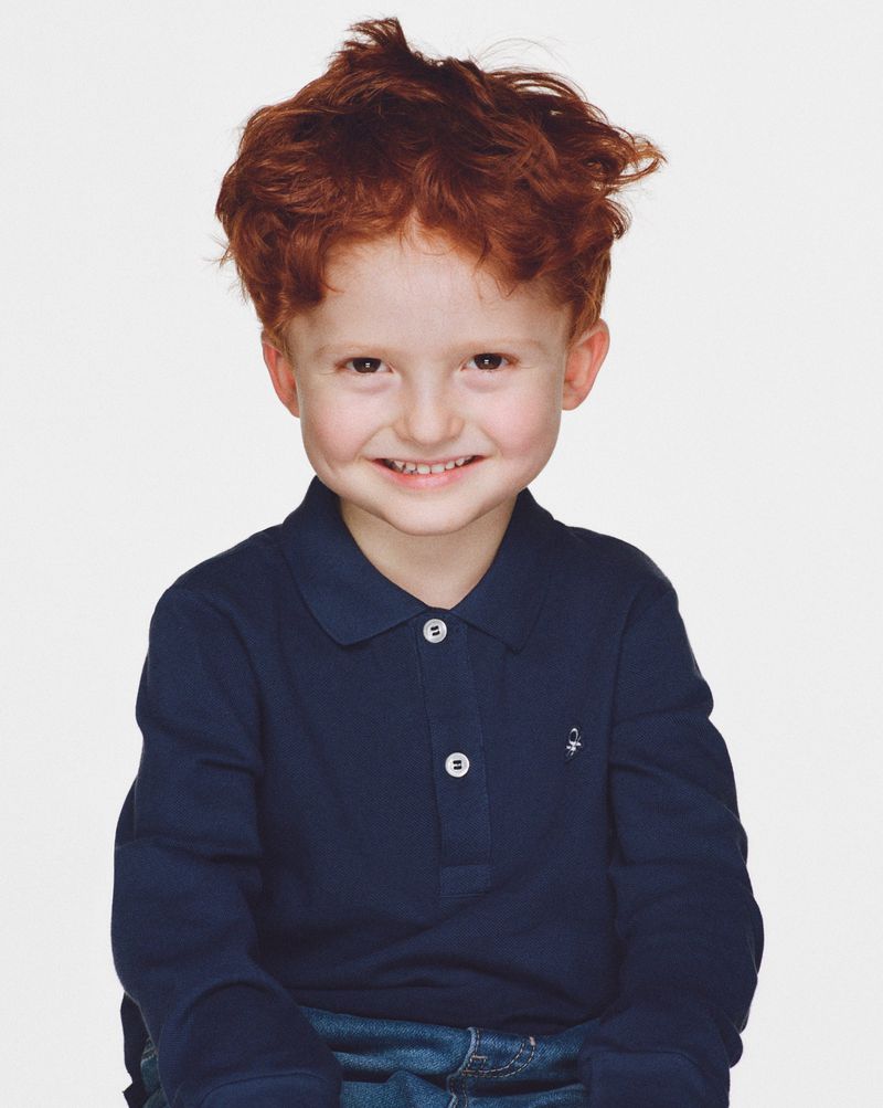 Portrait of a cute, smiling young boy with red hair, wearing a blue, long-sleeved polo shirt and jeans, against a white backdrop.