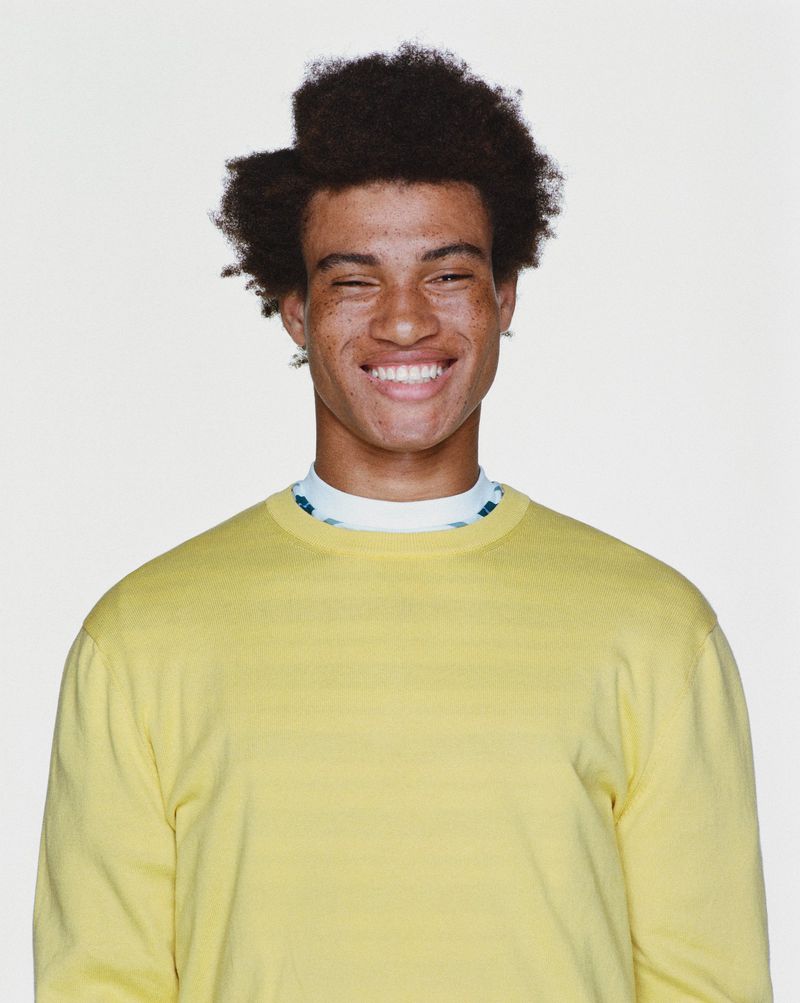 Portrait of a smiling young adult male with natural hair and a yellow sweatshirt on a white background, facing the camera.