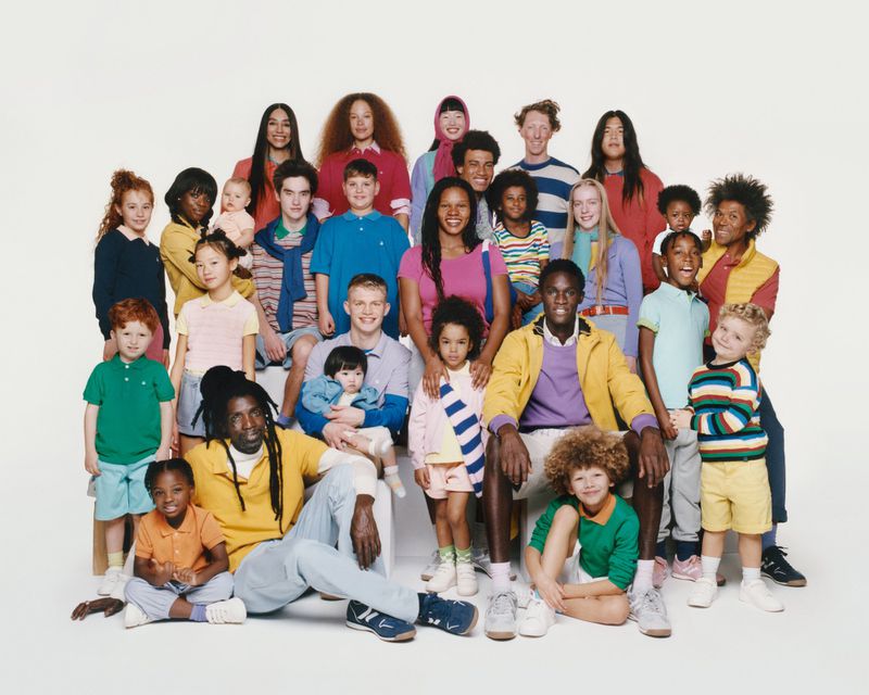 A diverse group portrait features twenty-six individuals of varying ages, ethnicities, and hairstyles, all dressed in casual clothing against a white backdrop.