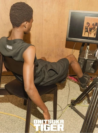 A Black man sits in a chair in an indoor setting, wearing an athletic outfit, with a computer monitor and camera equipment nearby. There is text at the bottom.