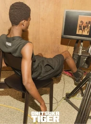 A Black male model wearing black athletic apparel seated on a dark chair looking at a monitor in an Onitsuka Tiger campaign shoot.