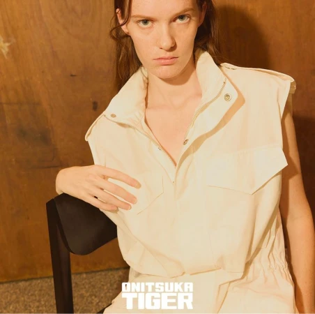 Model in sleeveless cream utility vest resting on a dark chair against a warm wooden wall