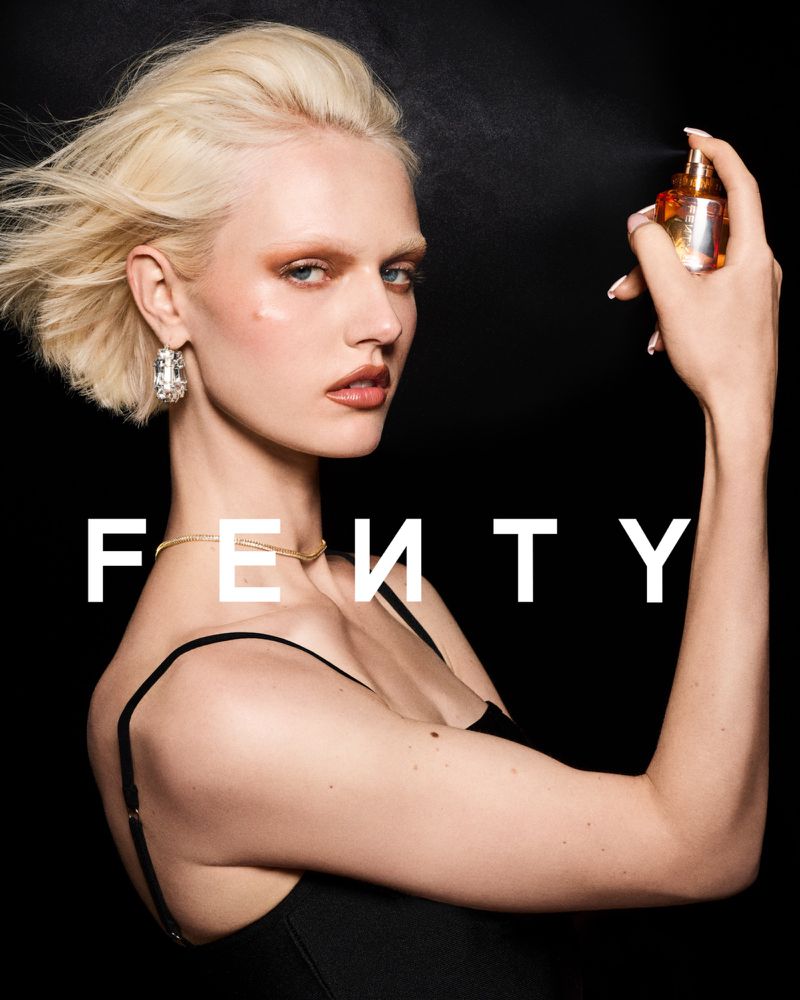 Woman with blonde hair applying perfume; fair skin, gold necklace, and large earrings; black tank top; black background, 'FENTY' text.