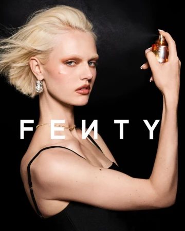 Woman with blonde hair applying perfume; fair skin, gold necklace, and large earrings; black tank top; black background, 'FENTY' text.