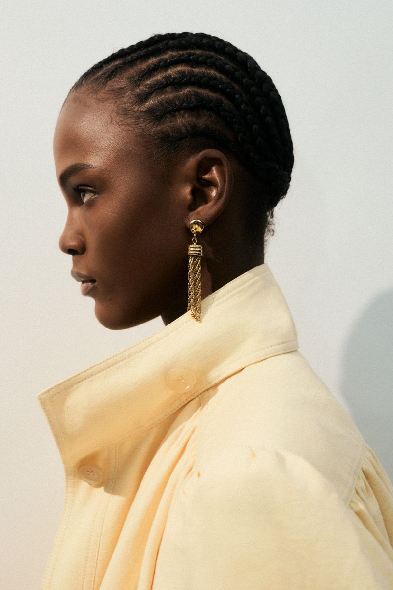 Profile portrait of a Black model in profile wearing a cream coat and gold tassel earrings against a white wall.