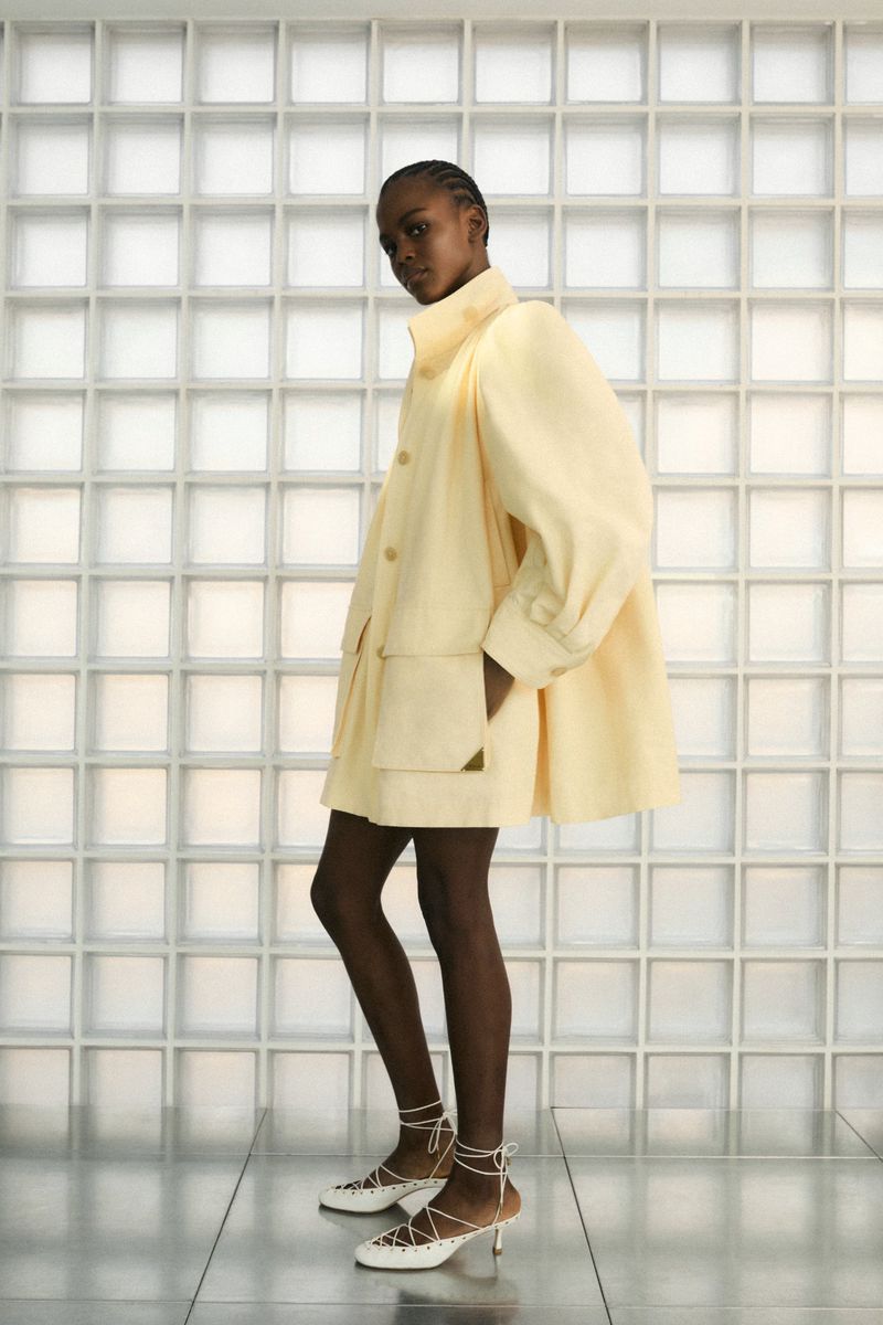 A Black female model showcases a light yellow jacket and strappy white heels, posing in a studio against a textured glass block wall.