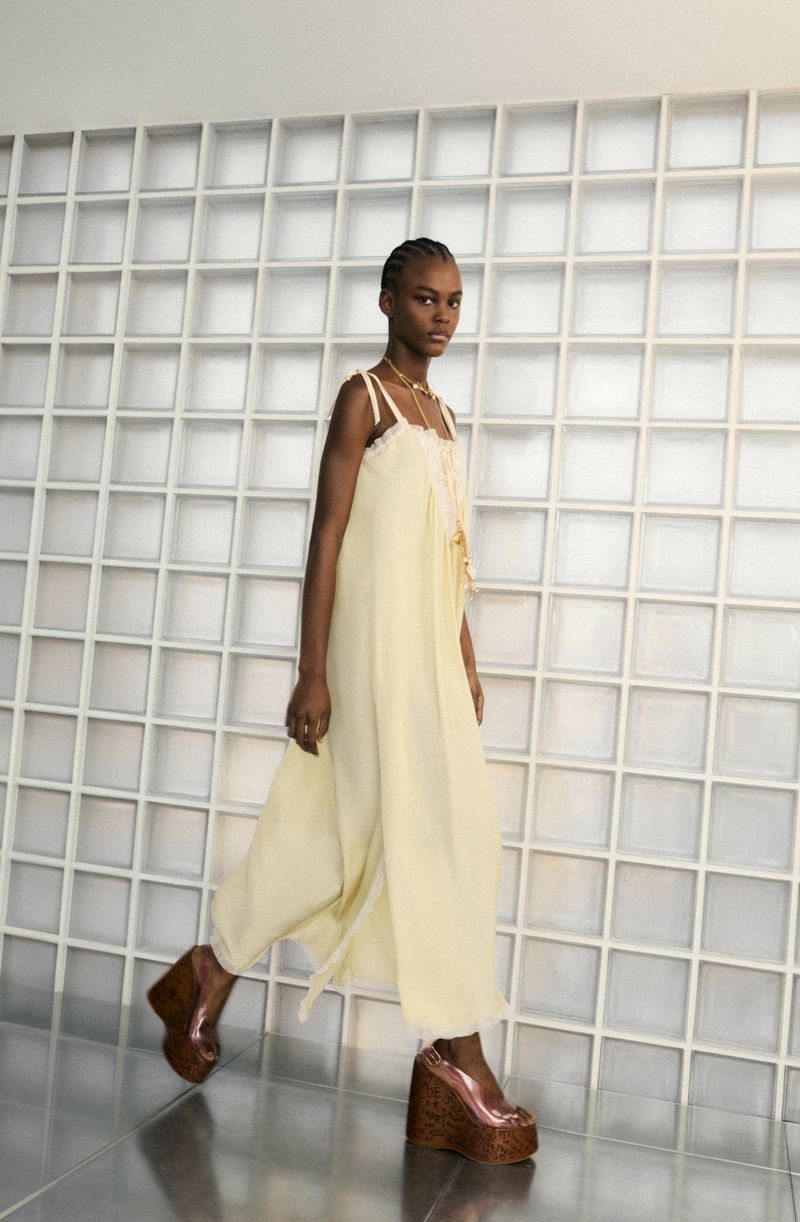 Fashion model in a pale yellow jumpsuit and platform heels walks in front of a white geometric wall, wearing a necklace.