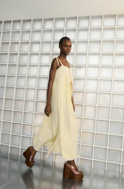 Fashion model in a pale yellow jumpsuit and platform heels walks in front of a white geometric wall, wearing a necklace.