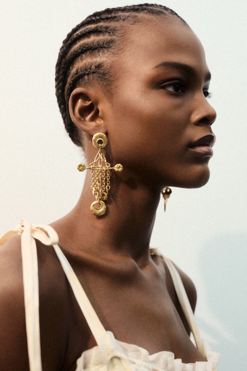 Close-up portrait of a Black model with braided hair wearing ornate gold drop earrings and a cream strap top against a white backdrop.