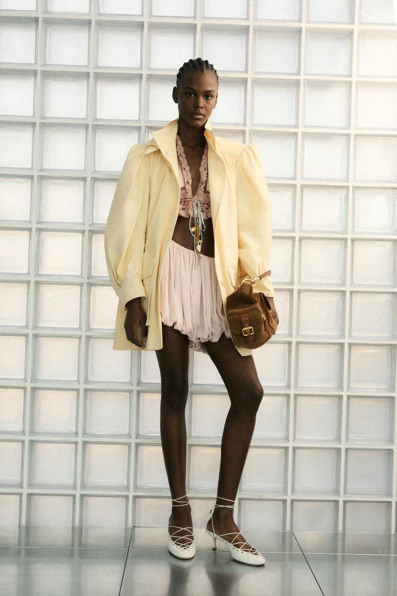 Fashion shot of a Black woman in a yellow jacket, pink shorts, and white shoes, carrying a brown purse against a white tiled wall.