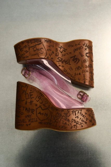 Platform wedge sandals with translucent straps and graffiti-style lettering in a still product shot showcasing fashion footwear.
