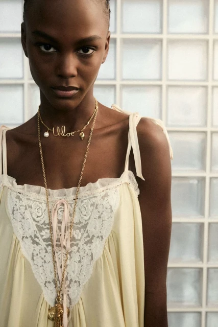 Close-up fashion shot of an African-American adult female model wearing a white lace-trimmed nightgown and Chloe logo pendant necklaces.
