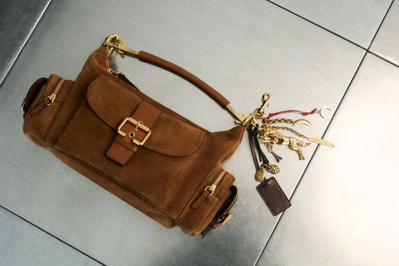 Brown suede hobo bag with gold hardware and multiple pockets, accessorized with playful charms, set against a neutral tiled background.