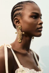 Close-up portrait of a Black model with braided hair wearing ornate gold drop earrings and a cream strap top against a white backdrop.