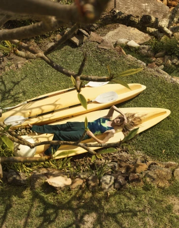 Model lying on a yellow kayak on grassy ground under tree branches for Bassike Resort 24