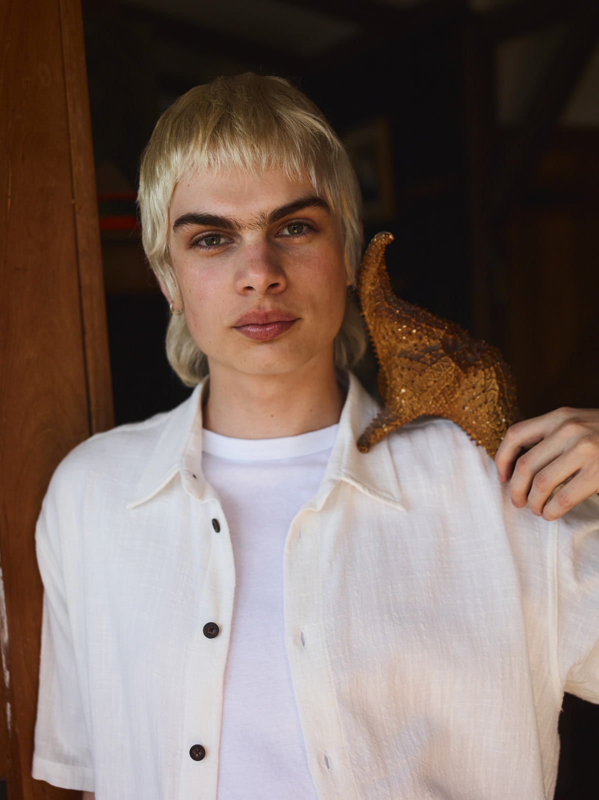Close-up portrait of a young adult with blonde hair, wearing a white shirt, holding a gold sequined object on their shoulder indoors.