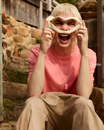 Blonde adult poses outdoors on stone steps, holding shark jawbone to face. Wears salmon-colored long-sleeve shirt and tan pants.