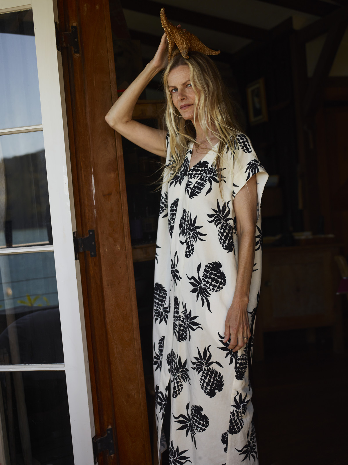 Elegant adult female in pineapple-print dress poses in doorway with starfish on head, bathed in natural light.