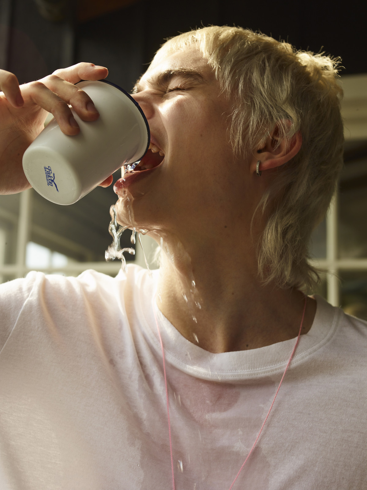 Close-up of a young adult with light hair drinking water from a white cup, with water spilling down their face and white shirt.