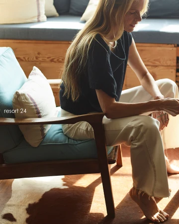 A woman with long blonde hair sits in a mid-century modern chair in a bright, airy room with a cowhide rug and sunlight streaming in.