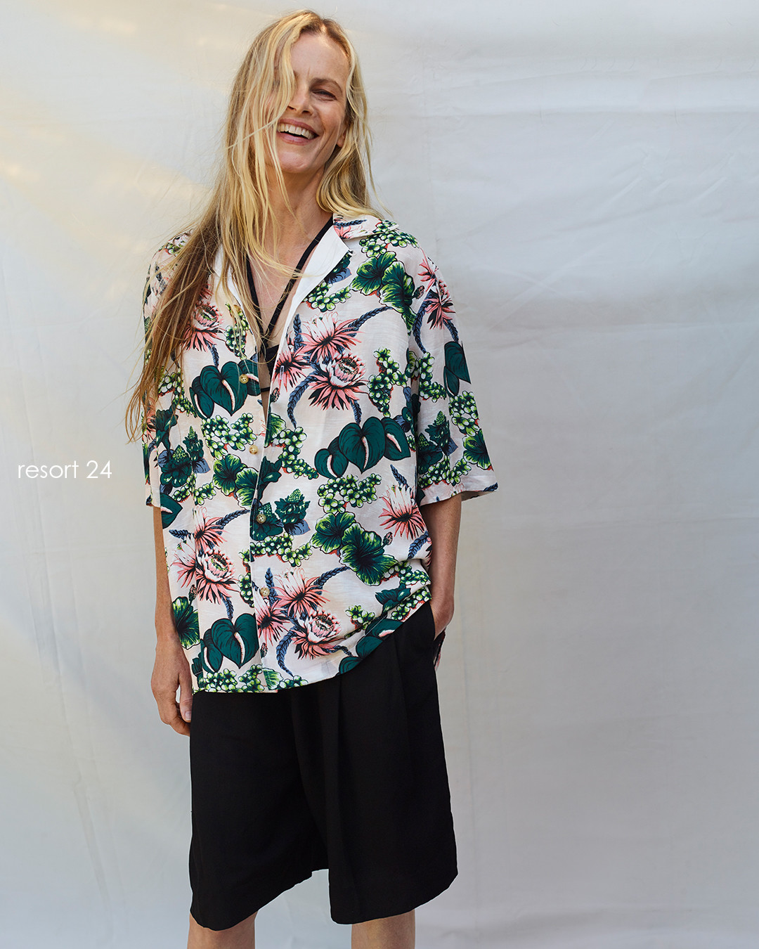 Smiling adult woman in a floral shirt with black shorts standing against a neutral backdrop. Fashionable resort wear.