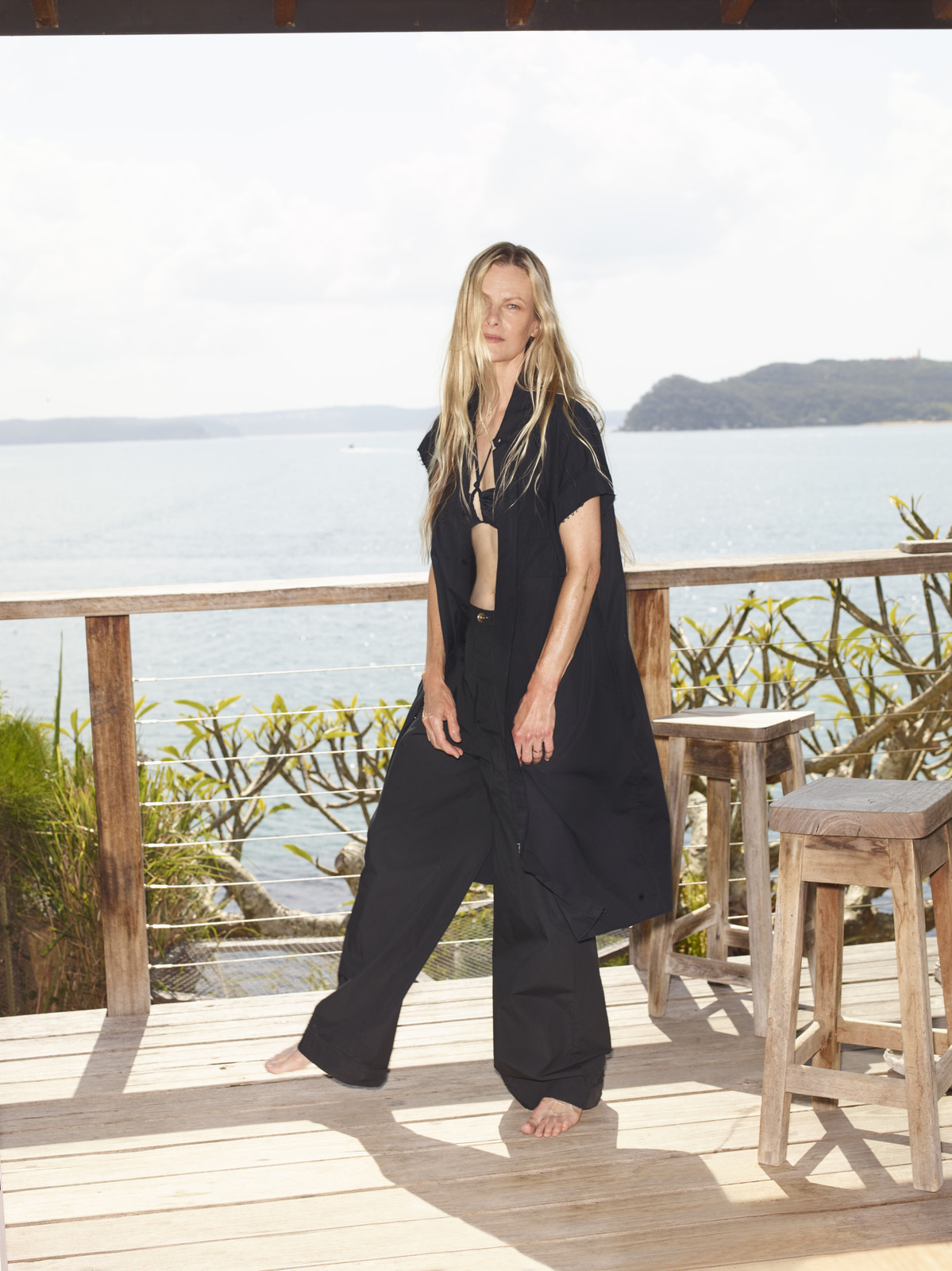 A barefoot, blonde woman in black casual clothing stands on a wooden deck overlooking the sea under a bright, sunny sky.
