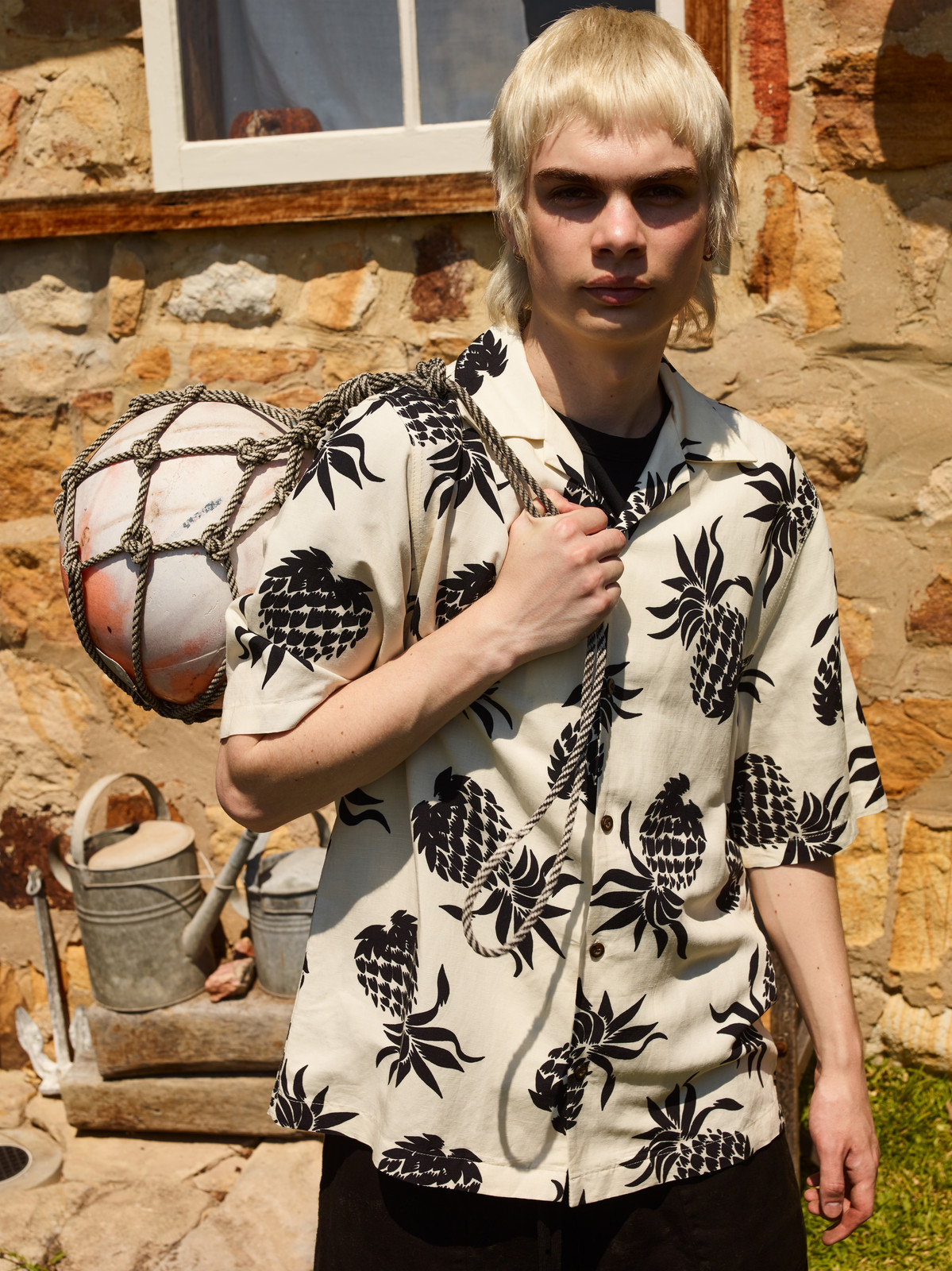 Male model poses outdoors in a pineapple-print shirt, carrying a rope-wrapped buoy over his shoulder, near a stone building with a window.