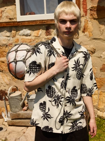Male model poses outdoors in a pineapple-print shirt, carrying a rope-wrapped buoy over his shoulder, near a stone building with a window.