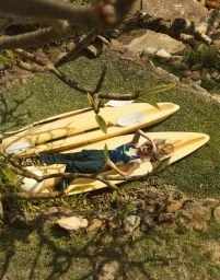 Model lying on a yellow kayak on grassy ground viewed through tree branches overhead