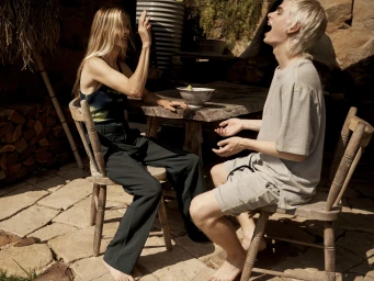 Two people seated outdoors at a wooden table on a stone patio, laughing and gesturing.