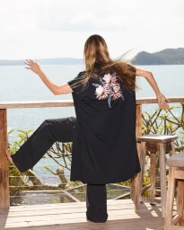Woman wearing black floral embroidered tunic and pants standing on a wooden balcony overlooking the ocean